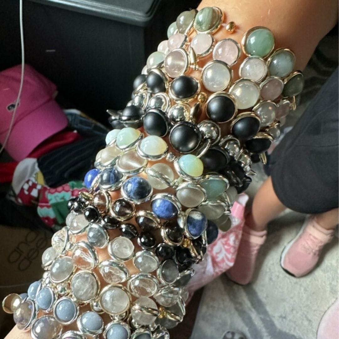 A close-up of a wrist wearing multiple stacked gemstone bracelets by Brett Lauren in various colors, shown outdoors with a casual outfit.