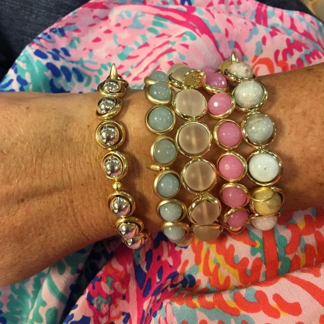 A close-up of a woman's wrist adorned with multiple Brett Lauren gemstone bracelets in pastel colors, including pink, grey, and gold accents.
